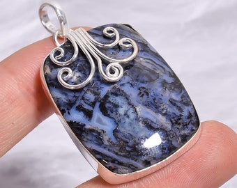 Puede incluir: Un colgante rectangular con una piedra pulida de patrones azules y negros engastada en un marco de plata. La parte superior del colgante está adornada con filigrana de plata en forma de espiral e incluye una reasa de plata para una cadena.