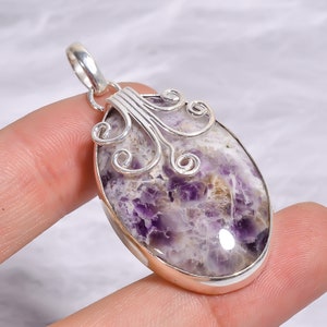 May include: An oval-shaped pendant featuring a polished amethyst stone with white and purple marbled patterns. The stone is set in a smooth silver bezel with decorative silver wire scrollwork at the top, including a silver bail for hanging.
