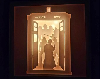 May include: A lit shadow box featuring a detailed silhouette of the iconic blue Police Box from the Doctor Who series. The box is open, revealing figures inside. The word "POLICE BOX" is visible on the top. The box is illuminated with a warm, inviting glow.