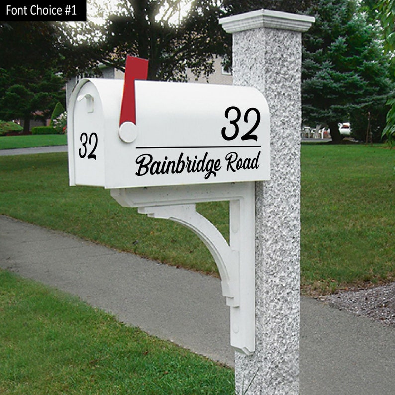 A white mailbox sit-in on a light white brick pole . The mailbox has a black mailbox decal that displays 32 Bainbridge Road on a cursive font. There is a small number 32 on the front lid of the mailbox.