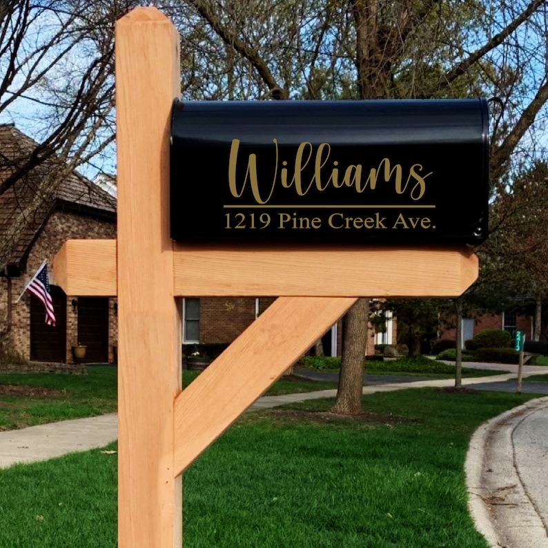 May include: A black mailbox with the name "Williams" in gold script and the address "1219 Pine Creek Ave." The mailbox is mounted on a natural wood post. The background includes a residential street and a house with an American flag.