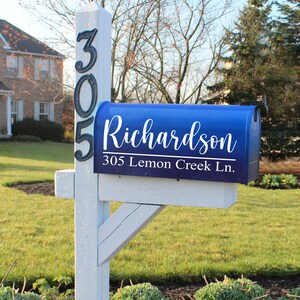 May include: A blue mailbox with the name "Richardson" and address "305 Lemon Creek Ln." in white script. The mailbox is mounted on a white wooden post with the numbers "305" in black. The scene is set in a residential area with a grassy lawn.