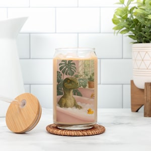 May include: A clear glass can-shaped cup with a bamboo lid and straw. The cup features a whimsical illustration of a green dinosaur relaxing in a pink bathtub with a yellow rubber ducky. The cup sits on a round, woven coaster.