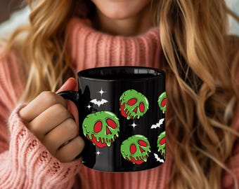 Puede incluir: Taza de cerámica negra con un patrón repetido de diseños de calaveras de manzana envenenada verdes, rojas y blancas. La taza también tiene gráficos de murciélagos y estrellas blancas. La taza es sostenida por una persona que lleva un suéter rosa.