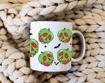 Puede incluir: Taza de cerámica blanca con un patrón repetido de manzanas verdes con forma de calavera con "veneno" rojo goteando y murciélagos negros. La taza tiene un asa blanca. Fondo crema tejido.