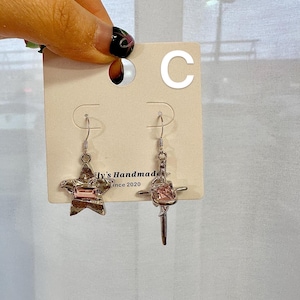 May include: A pair of silver earrings with pink crystal accents. One earring features a star shape and the other a cross shape. The earrings are displayed on a white card with the text "Lily's Handmade since 2020" and the letter "C" in the top right corner.
