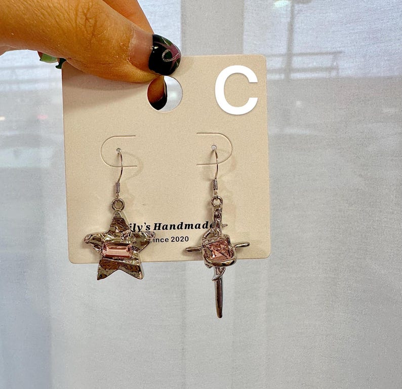 May include: A pair of silver earrings with pink crystal accents. One earring features a star shape and the other a cross shape. The earrings are displayed on a white card with the text "Lily's Handmade since 2020" and the letter "C" in the top right corner.