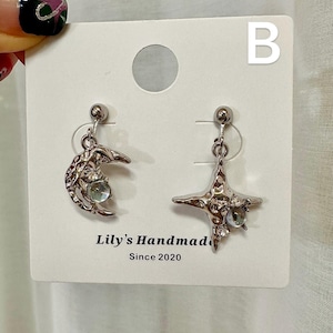 May include: A pair of silver earrings with a crescent moon and a star design. Each earring features a clear crystal stone. The earrings are displayed on a white card with the text "Lily's Handmade Since 2020" and the letter "B".