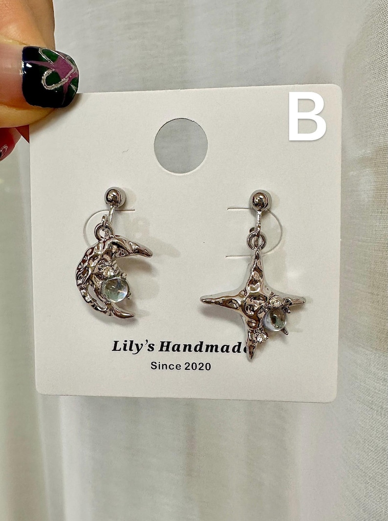 May include: A pair of silver earrings with a crescent moon and a star design. Each earring features a clear crystal stone. The earrings are displayed on a white card with the text "Lily's Handmade Since 2020" and the letter "B".