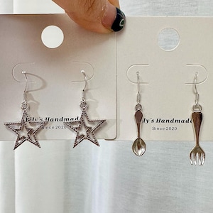 May include: Two pairs of silver earrings on white cards. The first pair features a star design. The second pair features a spoon and a fork design. The cards have the text "Billy's Handmade Since 2020" printed on them.
