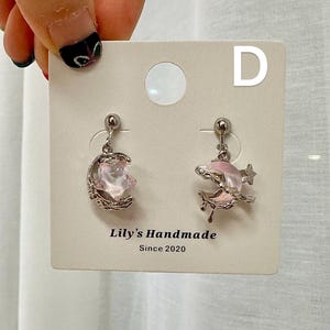 May include: A pair of silver earrings with pink crescent moon and star charms. The earrings are displayed on a white card with the text "Lily's Handmade Since 2020" and the letter "D".