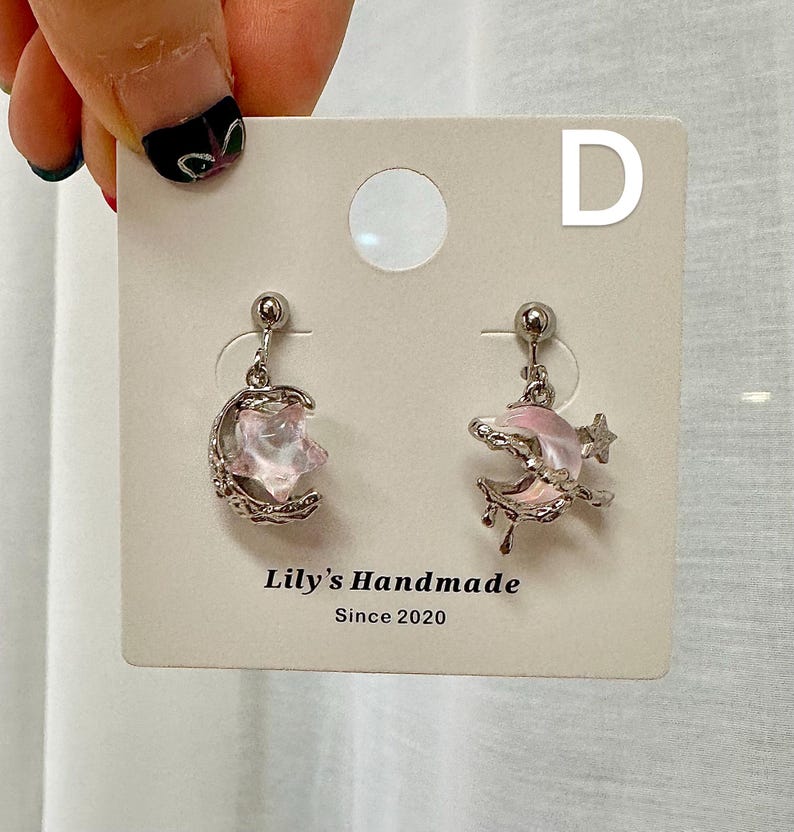 May include: A pair of silver earrings with pink crescent moon and star charms. The earrings are displayed on a white card with the text "Lily's Handmade Since 2020" and the letter "D".