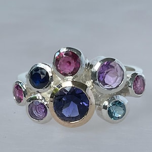 May include: A silver ring with a cluster of colourful gemstones. The ring features a large, deep purple centre stone with a gold bezel, surrounded by smaller gems in shades of pink, blue, and purple. The ring's design is unique and eye-catching.