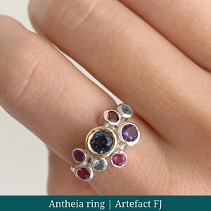 May include: A silver ring with a cluster of colourful gemstones. The ring features a large, dark blue centre stone surrounded by smaller gems in shades of purple, pink, and blue. The ring has a gold band around the centre stone. Text on the image reads: Antheia ring | Artefact FJ.
