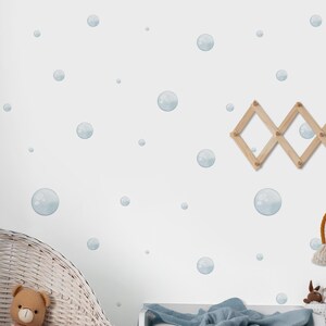 May include: A nursery scene with a white wall adorned with blue bubble decals. A wicker bassinet holds a teddy bear and blanket. A wooden accordion peg rack hangs a rainbow-shaped wall hanging. A changing table is also present.