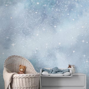 Pode incluir: Uma cena de berçário com uma parede em aquarela azul clara com estrelas brancas. Um berço de vime branco contém um ursinho de pelúcia e um cobertor. Uma mesa de mudança com um cobertor azul e uma pequena cesta com brinquedos completam a cena.