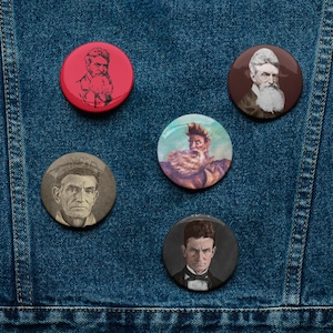 May include: Five round pinback buttons with portraits of historical figures. The buttons are red, brown, and gray, with images of men with beards and formal attire. The buttons are arranged on a blue denim background.