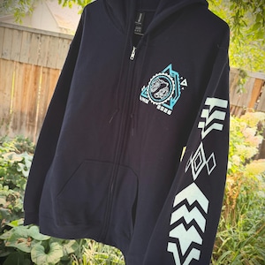 May include: A dark navy blue zip-up hoodie featuring a light blue graphic on the left chest with a circular design, the text "ANU 2026," and a geometric pattern. The left sleeve displays a series of light blue chevron and diamond shapes.