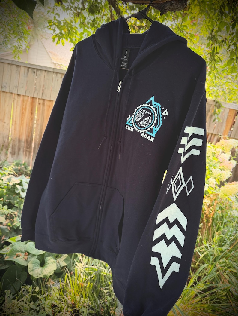 May include: A dark navy blue zip-up hoodie featuring a light blue graphic on the left chest with a circular design, the text "ANU 2026," and a geometric pattern. The left sleeve displays a series of light blue chevron and diamond shapes.