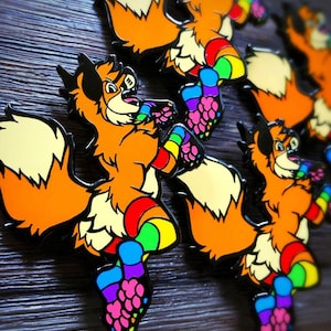 May include: A set of four enamel pins featuring a cartoon fox wearing rainbow-colored clothing. The fox has a fluffy tail and is depicted in a playful pose. The pins are a vibrant orange, yellow, green, blue, and purple.