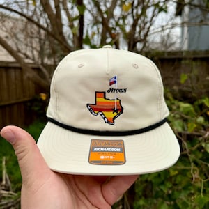 Könnte beinhalten: Eine weiße Baseballkappe mit schwarzem Schirm und einem Seildetail. Die Kappe zeigt ein gesticktes Bild des Bundesstaates Texas mit einer Flagge auf einem Golf-Fahnenmast. Der Text "ATOWN" ist über dem Bild gestickt. Die Kappe hat ein Richardson R-Active-Etikett mit einem Lichtschutzfaktor UPF 50+.