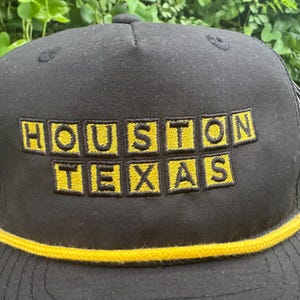 Może przedstawiać: Czarna czapka bejsbolowa z haftowanym napisem "HOUSTON TEXAS" w ż&oacute;łtych literach blokowych. Czapka ma ż&oacute;łty sznurek wzdłuż ronda.