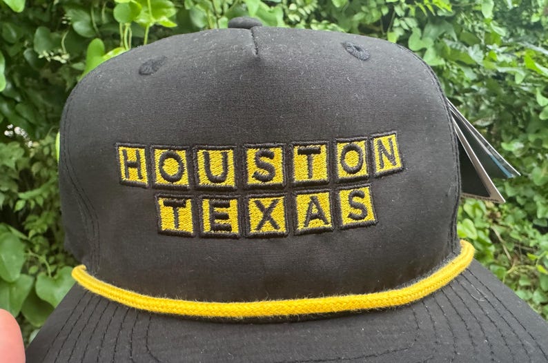 Może przedstawiać: Czarna czapka bejsbolowa z haftowanym napisem "HOUSTON TEXAS" w ż&oacute;łtych literach blokowych. Czapka ma ż&oacute;łty sznurek wzdłuż ronda.