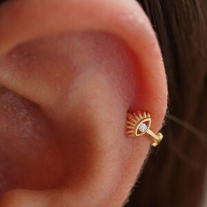 May include: A gold-colored earring in the shape of an eye, with a small, clear gem in the center. The earring is attached to the ear with a small hoop. The design features sun rays around the eye, creating a unique and stylish look. Jewelry.