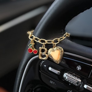 May include: Gold-toned charm bracelet with a cherry, letter "B", and heart pendant, hanging from a black steering wheel. The cherry is red with green leaves. The heart pendant has a gold frame and a yellow center.