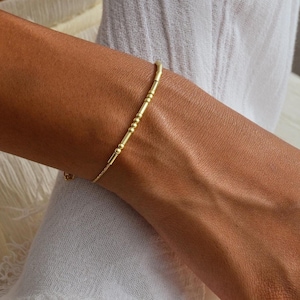 May include: A delicate gold bracelet with a linear design is displayed. The bracelet features a series of small, evenly spaced segments and is worn on a wrist. The image is set against a backdrop of white fabric.