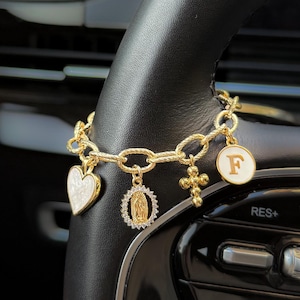 May include: Gold-toned charm bracelet with multiple pendants, including a heart, a religious figure, a cross, and a circular charm with the letter "F". The bracelet is displayed on a black steering wheel.