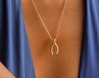 May include: A gold-colored wishbone pendant necklace. The pendant is suspended from a delicate chain. The necklace is worn against a blue fabric background.
