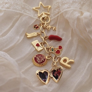 Puede incluir: Pulsera de dijes dorada con varios dijes, incluyendo una estrella, una llave, una mariquita, un sobre, un chile, cerezas, un relicario en forma de coraz&oacute;n y la letra "R". Los dijes cuelgan de una cadena sobre una tela color crema.