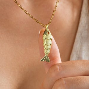 May include: A gold-colored fish pendant necklace. The fish has detailed scales and a small red accent. The necklace chain is also gold and has a delicate, rectangular link design. The necklace is being held up by a person.