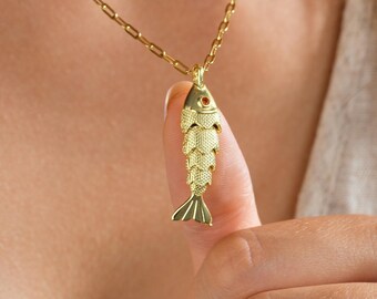 May include: A gold-colored fish pendant necklace. The fish has detailed scales and a small red accent. The necklace chain is also gold and has a delicate, rectangular link design. The necklace is being held up by a person.