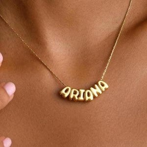 May include: A gold-colored necklace with the name "Ariana" spelled out in individual, rounded letters. The necklace is worn around the neck, with the chain and name pendant visible against the skin. The person is wearing a white top.