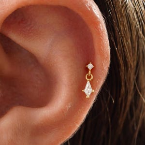 May include: A close-up of an ear with a small, gold earring. The earring features a small, round diamond stud and a dangling, teardrop-shaped diamond. The jewelry is delicate and elegant.