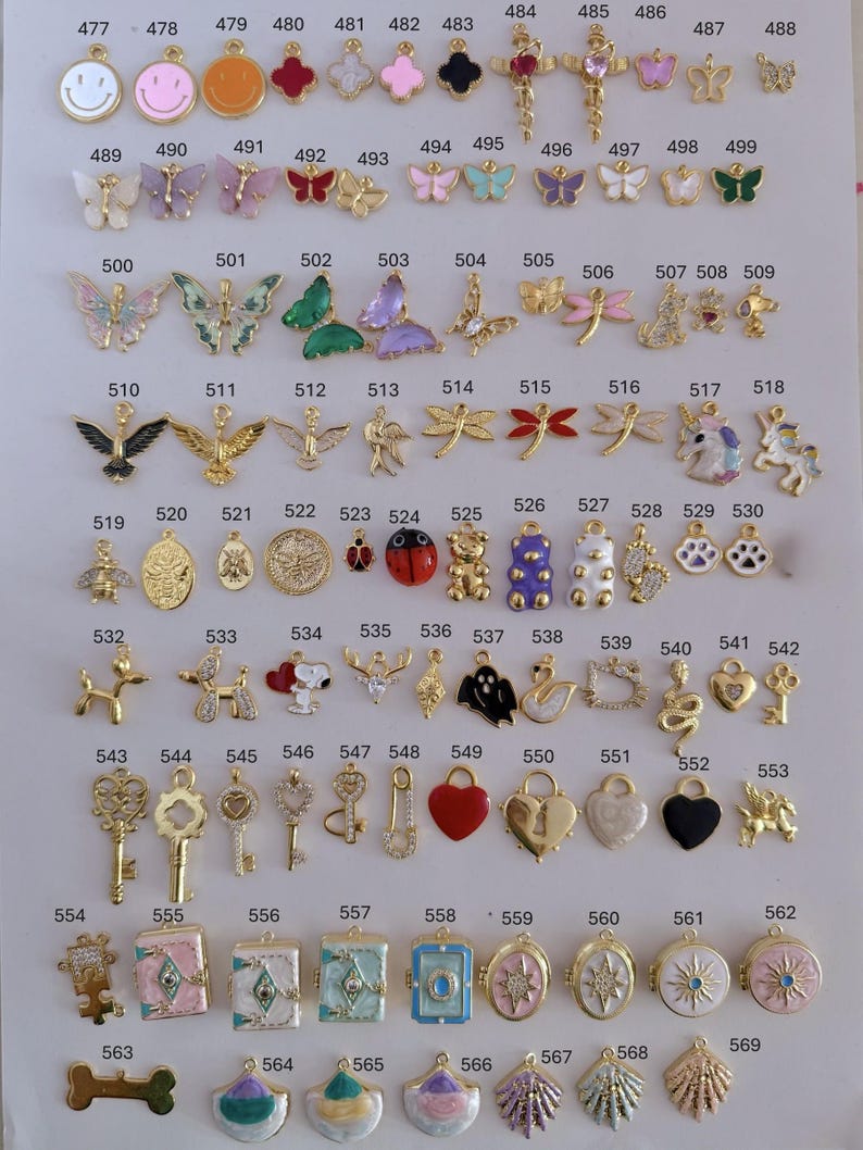 Puede incluir: Una colecci&oacute;n de colgantes dorados en varias formas, incluyendo mariposas, llaves, corazones y animales. Los colgantes est&aacute;n dispuestos sobre una superficie blanca, cada uno etiquetado con un n&uacute;mero. Adecuado para la creaci&oacute;n de joyas.