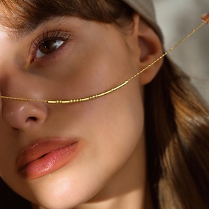 May include: A close-up of a gold-colored necklace with a series of small, cylindrical beads. The necklace is draped across the model's face, highlighting the delicate design. The model has brown eyes and is wearing a neutral-toned head covering.