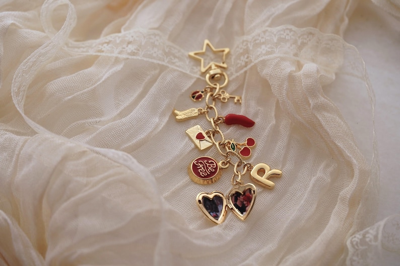 Puede incluir: Pulsera de dijes dorada con varios dijes, incluyendo una estrella, una llave, una mariquita, un sobre, un chile, cerezas, un relicario en forma de coraz&oacute;n y la letra "R". Los dijes cuelgan de una cadena sobre una tela color crema.