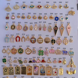 Puede incluir: Una colecci&oacute;n de colgantes y dijes dorados, con varios dise&ntilde;os como ojos turcos, manos de F&aacute;tima, tr&eacute;boles de cuatro hojas, elefantes y motivos de cartas del tarot. Los dijes est&aacute;n dispuestos sobre una superficie blanca.
