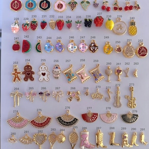 Puede incluir: Una colecci&oacute;n de abalorios dorados con diversos dise&ntilde;os, incluyendo frutas, postres, instrumentos musicales y lazos. Los abalorios est&aacute;n dispuestos sobre una superficie blanca, mostrando sus intrincados detalles y variados temas.