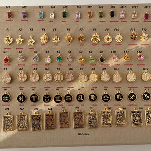 Puede incluir: Una colecci&oacute;n de colgantes y dijes dorados, que incluyen signos del zodiaco, letras, piedras de nacimiento y dise&ntilde;os de cartas del tarot. Los dijes est&aacute;n dispuestos sobre una superficie beige, con etiquetas de texto para cada art&iacute;culo.