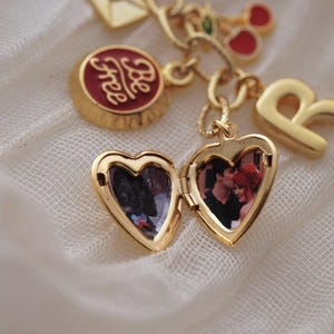 Puede incluir: Una pulsera de dijes dorada con un relicario en forma de coraz&oacute;n, un dije redondo con la inscripci&oacute;n "Be free", una letra "R" y otros dijes decorativos. El relicario est&aacute; abierto, revelando una fotograf&iacute;a.