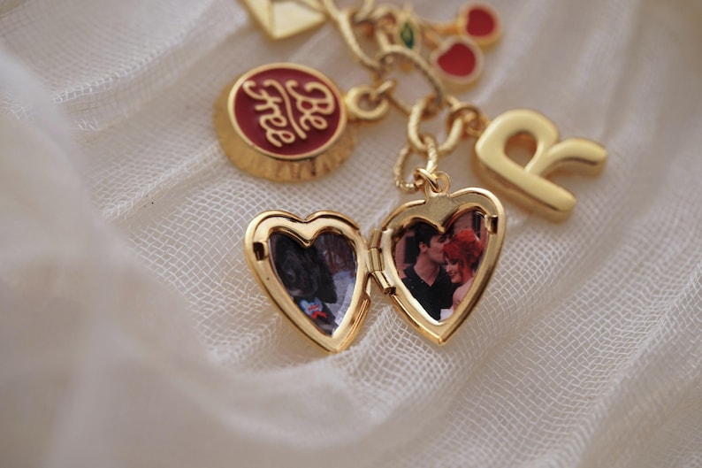 Puede incluir: Una pulsera de dijes dorada con un relicario en forma de coraz&oacute;n, un dije redondo con la inscripci&oacute;n "Be free", una letra "R" y otros dijes decorativos. El relicario est&aacute; abierto, revelando una fotograf&iacute;a.
