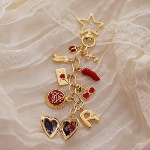 Puede incluir: Una pulsera de dijes dorada con varios dijes, incluyendo una estrella, una llave, un relicario en forma de coraz&oacute;n y un dije que dice "Be Free". Otros dijes incluyen un chile, cerezas y la letra "R".