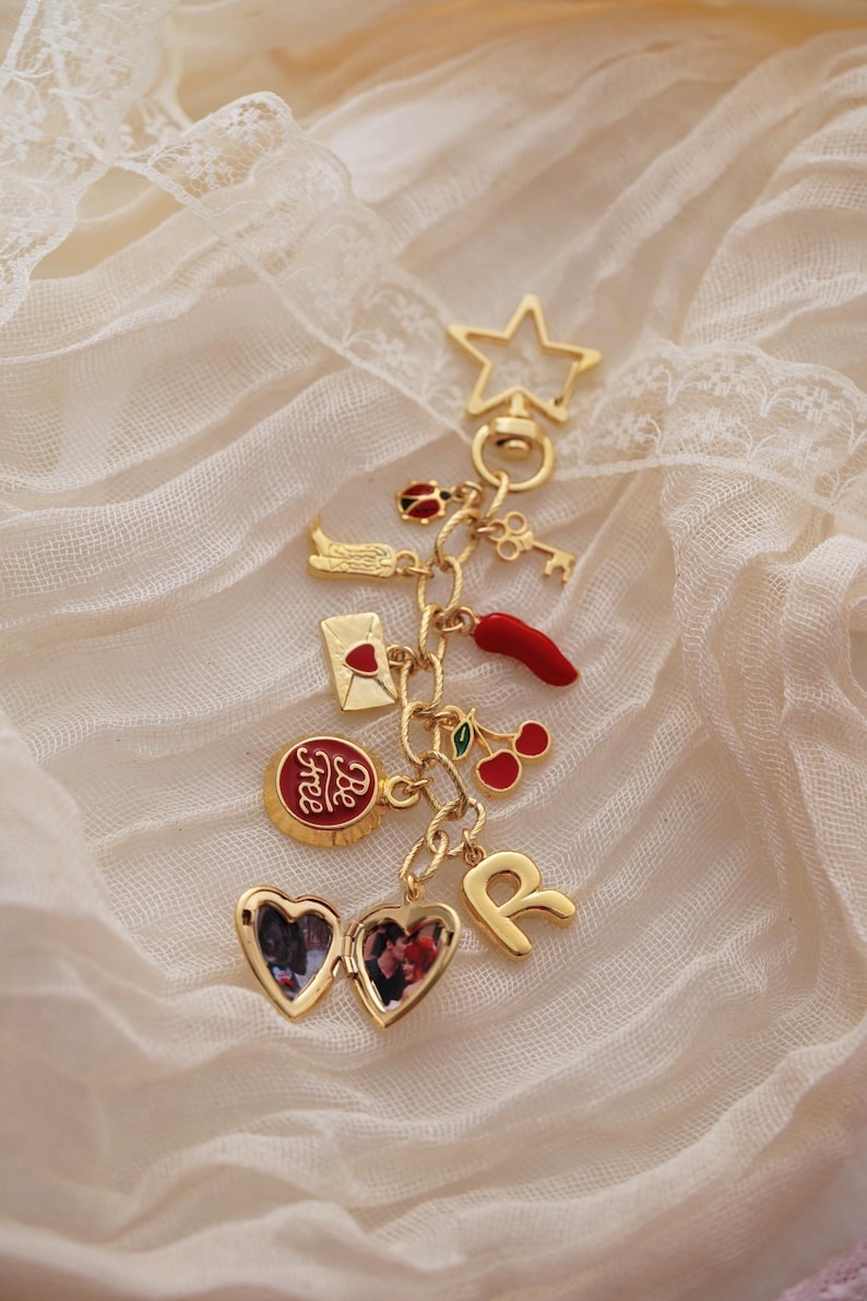 Puede incluir: Una pulsera de dijes dorada con varios dijes, incluyendo una estrella, una llave, un relicario en forma de coraz&oacute;n y un dije que dice "Be Free". Otros dijes incluyen un chile, cerezas y la letra "R".