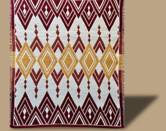 Puede incluir: Una manta de ganchillo hecha a mano con un patrón geométrico de diamantes en tonos burdeos, blanco y dorado. La manta presenta un diseño repetitivo de formas de diamantes y triángulos, con un borde burdeos. El texto en la parte inferior dice "Gemini Ember Test by: Andrea Aggarwal."