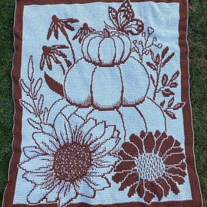 Puede incluir: Una manta de ganchillo con un diseño marrón y blanco con calabazas, girasoles y mariposas. La manta tiene un fondo azul claro y un borde marrón.