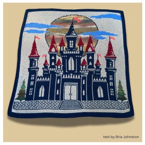 May include: A navy blue and white crocheted blanket featuring a castle design. The castle has red-topped towers, white windows, and a large sun in the background. The blanket has a navy blue border and the text "test by Bria Johnston".